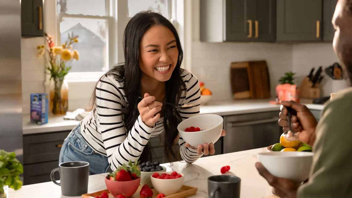 A woman smiling and enjoying breakfast with fresh berries and cereals at a modern kitchen table.
